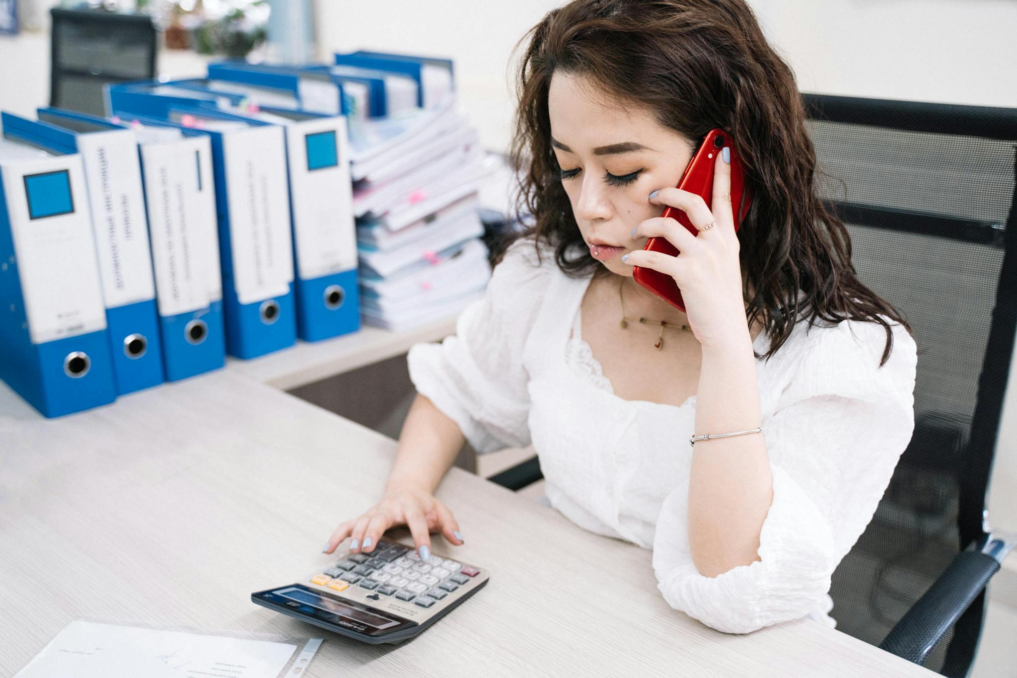 Businesswoman multitasking with calculator and cell phone at office desk.